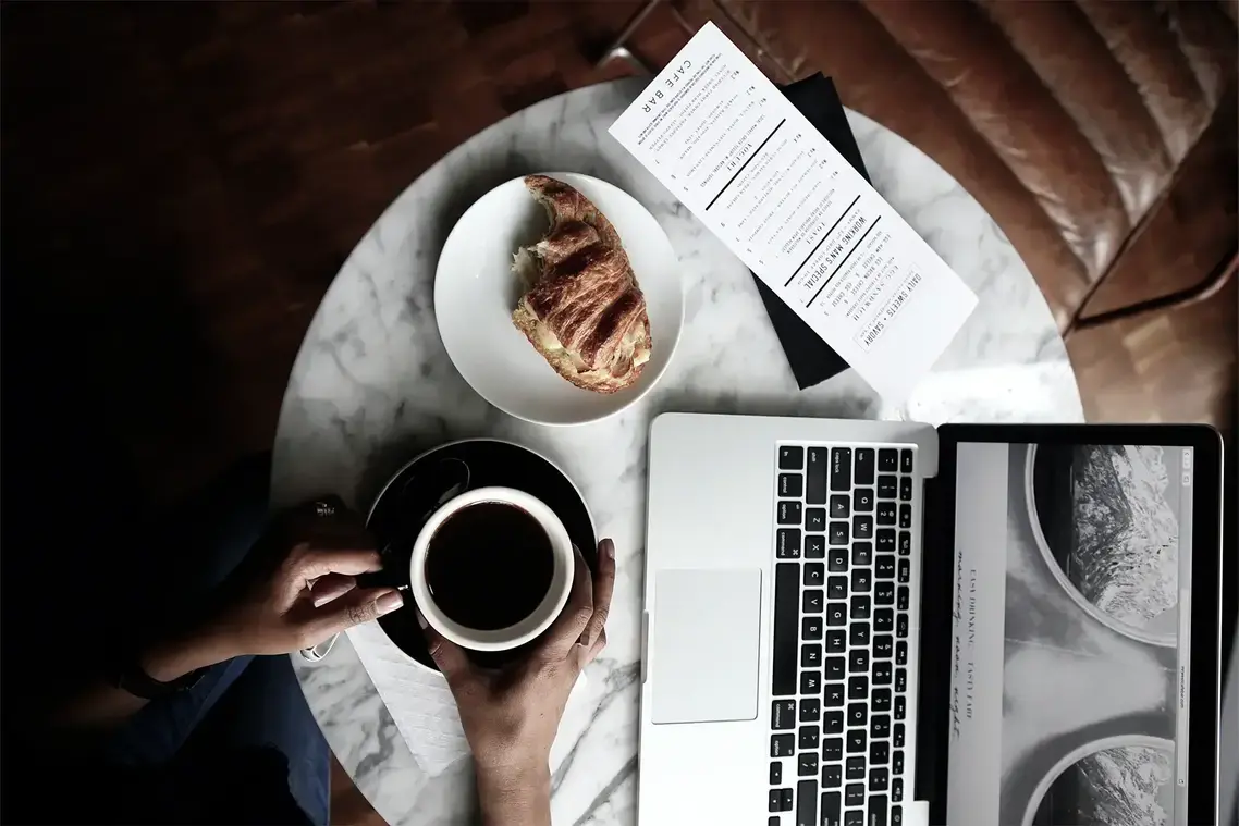 Tavolino da bar con tazza di caffè e croissant, sfogliando la schermata di un notebook