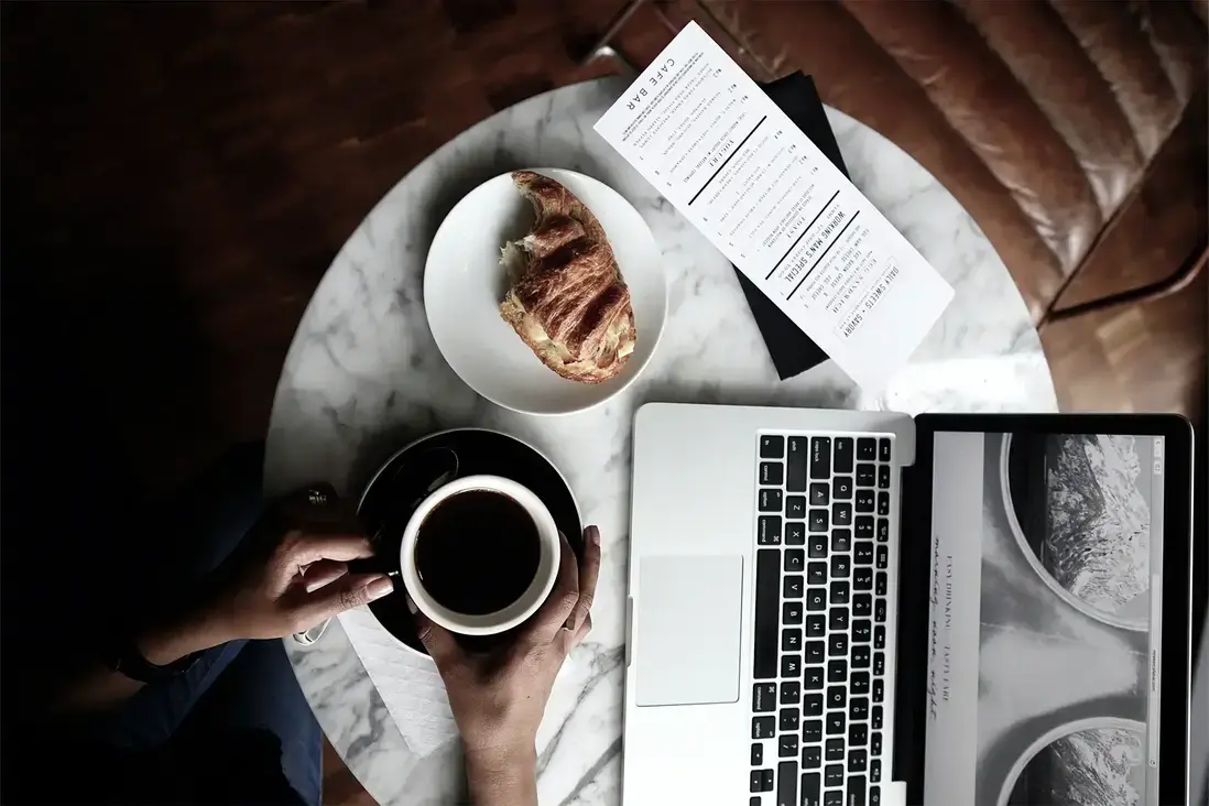 Tavolino da bar con tazza di caffè e croissant, sfogliando la schermata di un notebook
